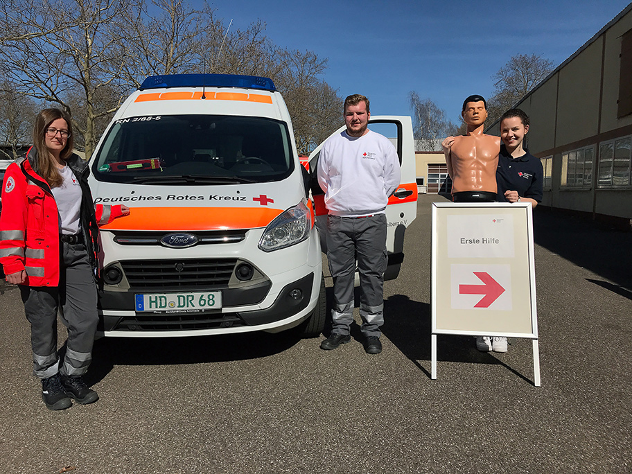 Drei DRK-Mitarbeiter neben einem Einsatzfahrzeug und Schild Erste Hilfe