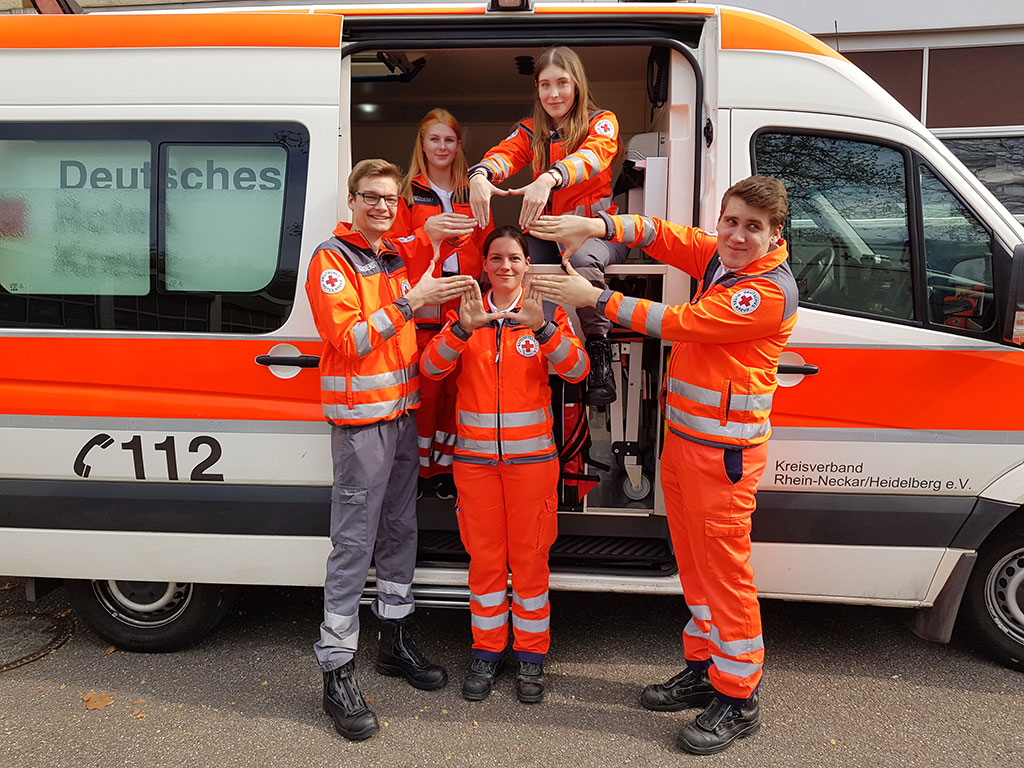 5 NotSan-Schüler*innen stehen vor einem Rettungswagen und bilden mit ihren Händen ein Kreuz
