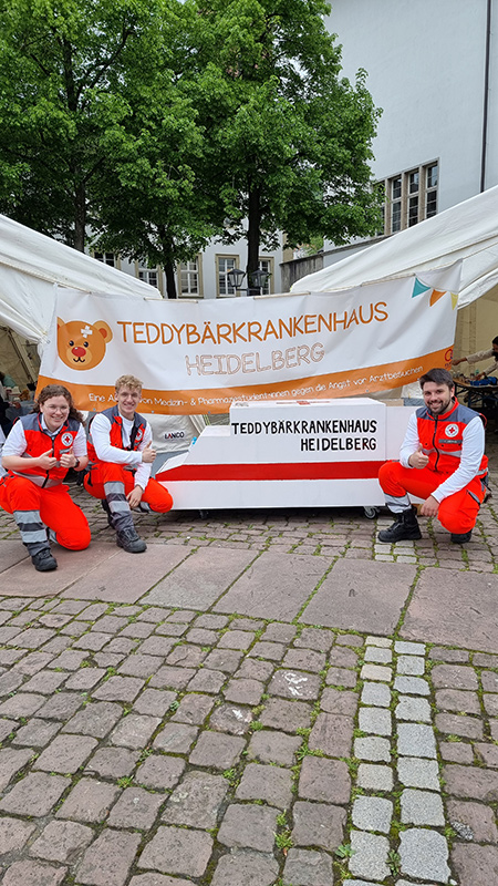 Drei DRK Notfallsanitäter/innen vor einem Banner mit der Aufschrift Teddybärenkrankenhaus Heidelberg