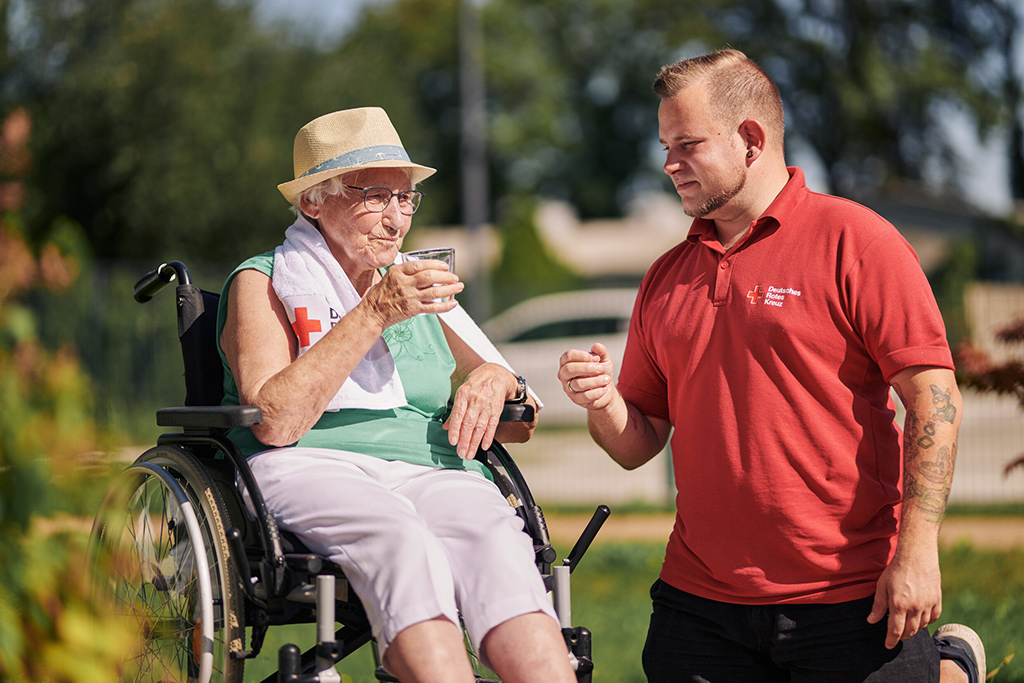 Überhitzte Seniorin im Rollstuhl erhält von einem DRK-Mitarbeiter ein Glas Wasser